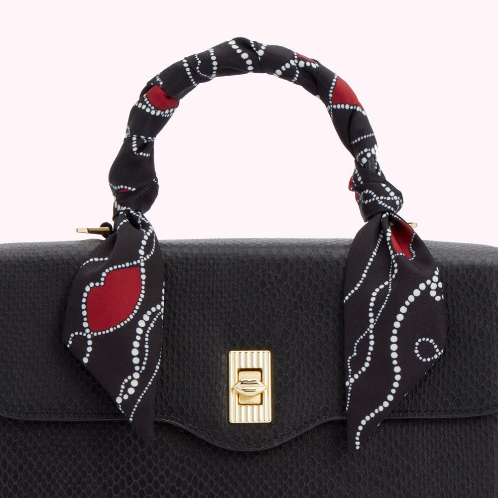 Close-up of a black handbag with a black and red patterned scarf tied around the handle. The background is white.
