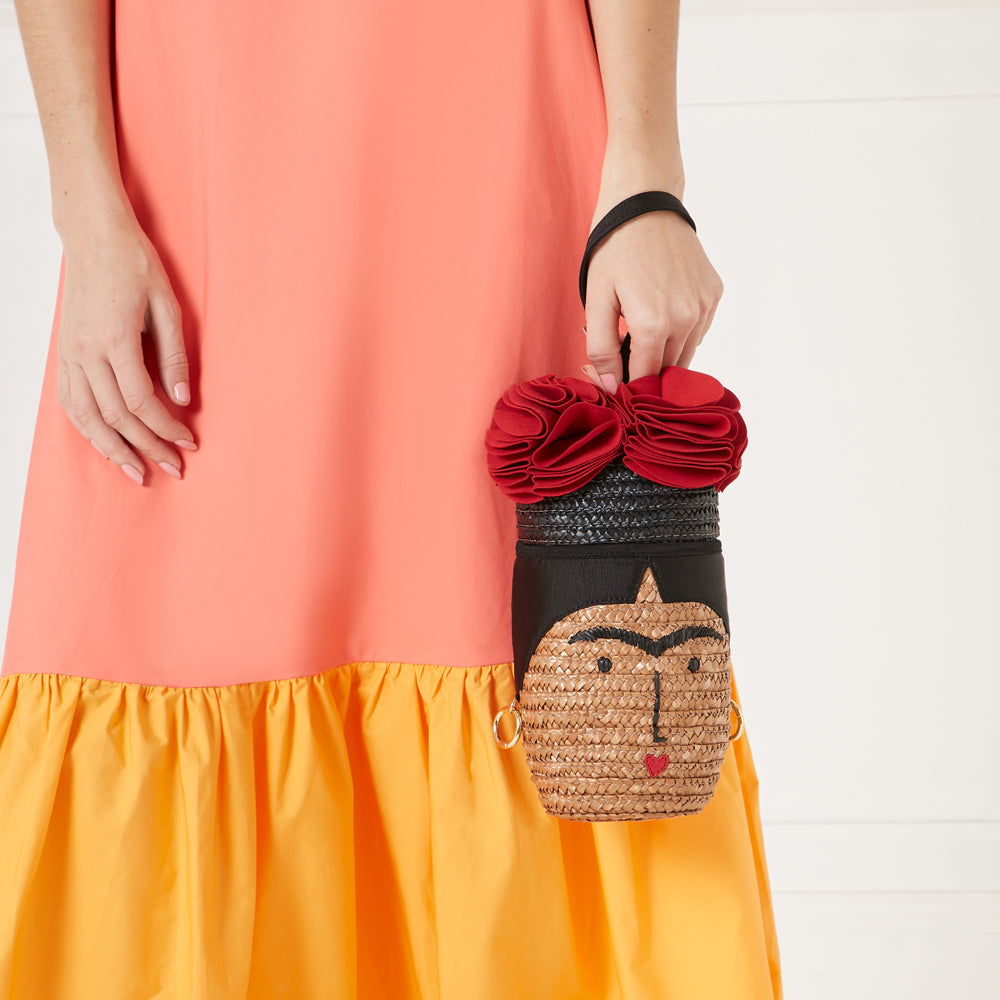 Woman in coral and yellow dress holding a Frida Kahlo-inspired straw handbag with red flower detail against a white background.
