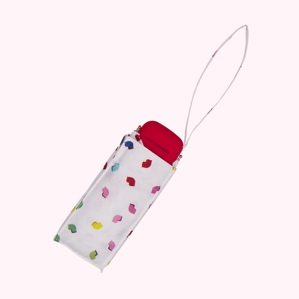 Compact Lulu Guinness umbrella in its case; white case with multicolored lips print; red umbrella handle.
