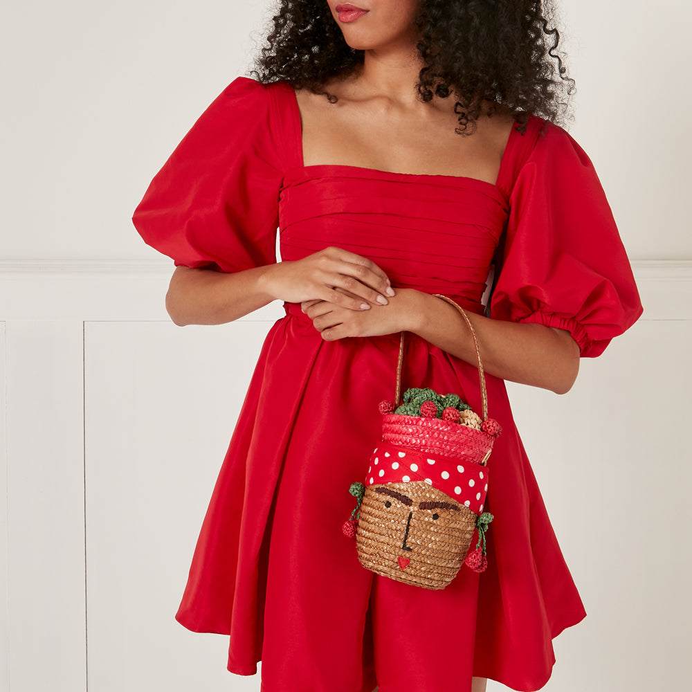 Woman in red dress holding a straw handbag shaped like a face with fruit on top.