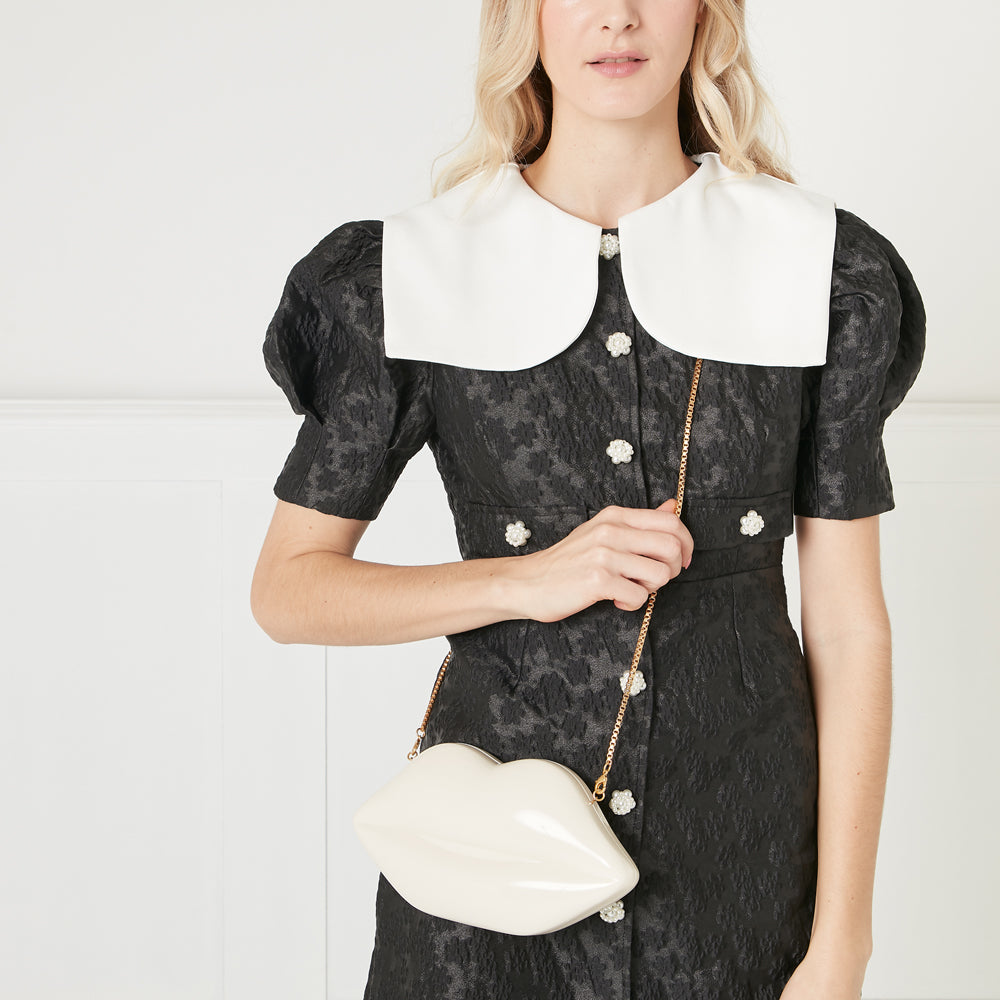 Woman in black dress with large white collar holding off-white lip-shaped bag.
