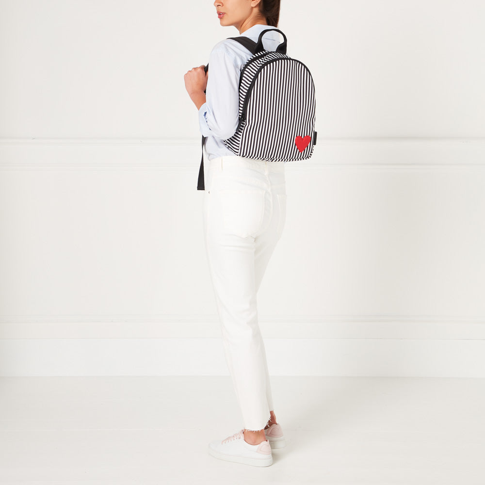 Woman wearing white jeans and a light blue shirt, seen from behind, carrying a black and white striped backpack with a small red heart detail. The background is white.
