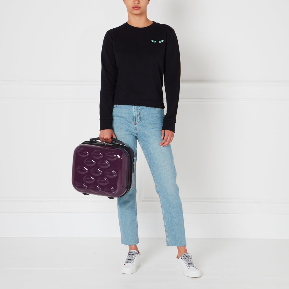 A woman in light blue jeans and a black sweater holds a purple carry-on suitcase with a lip print design against a white background.