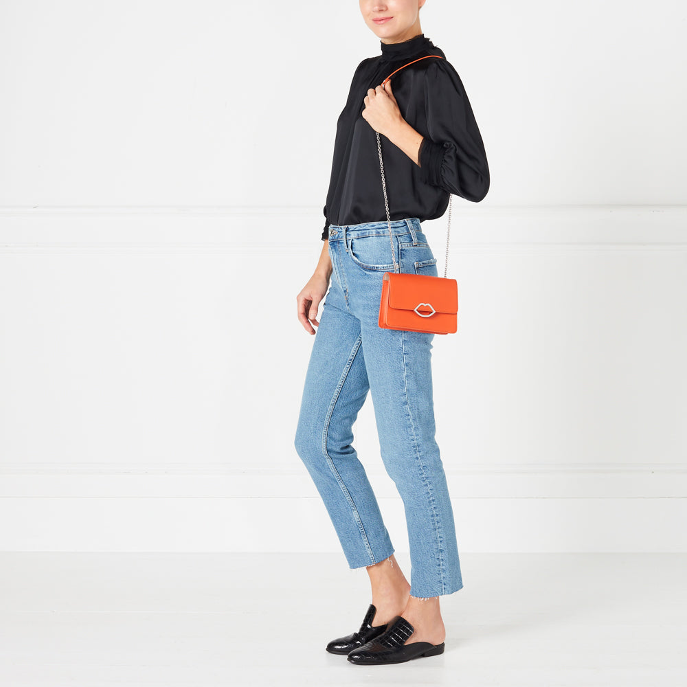 Woman in black top and light wash jeans wearing orange lip-shaped crossbody bag and black shoes against a white background.
