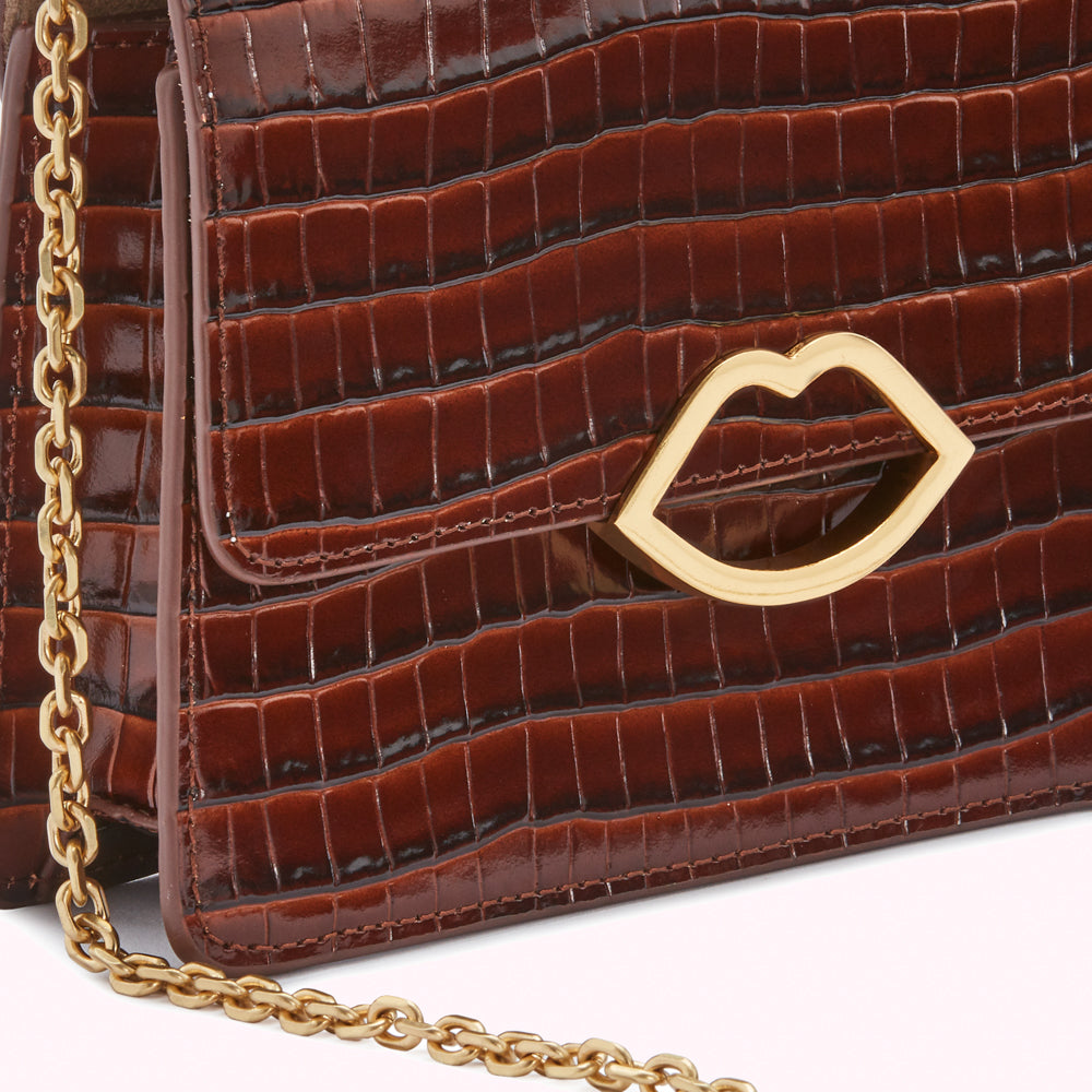 Close-up of a brown crocodile-embossed leather handbag with a gold lip-shaped clasp and gold chain strap.
