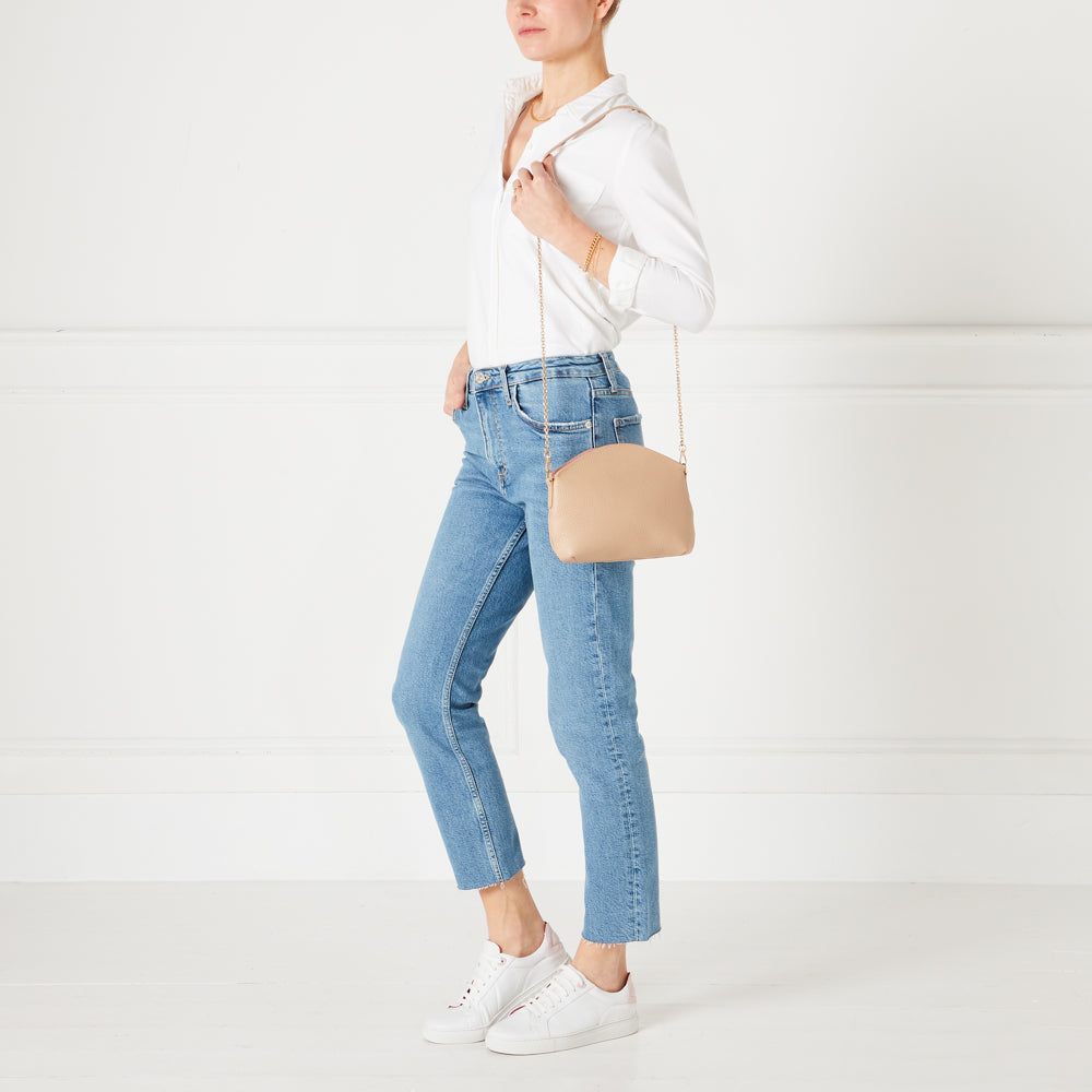 A woman in a white shirt and jeans models a light beige crossbody bag with a rose gold chain strap against a white background.
