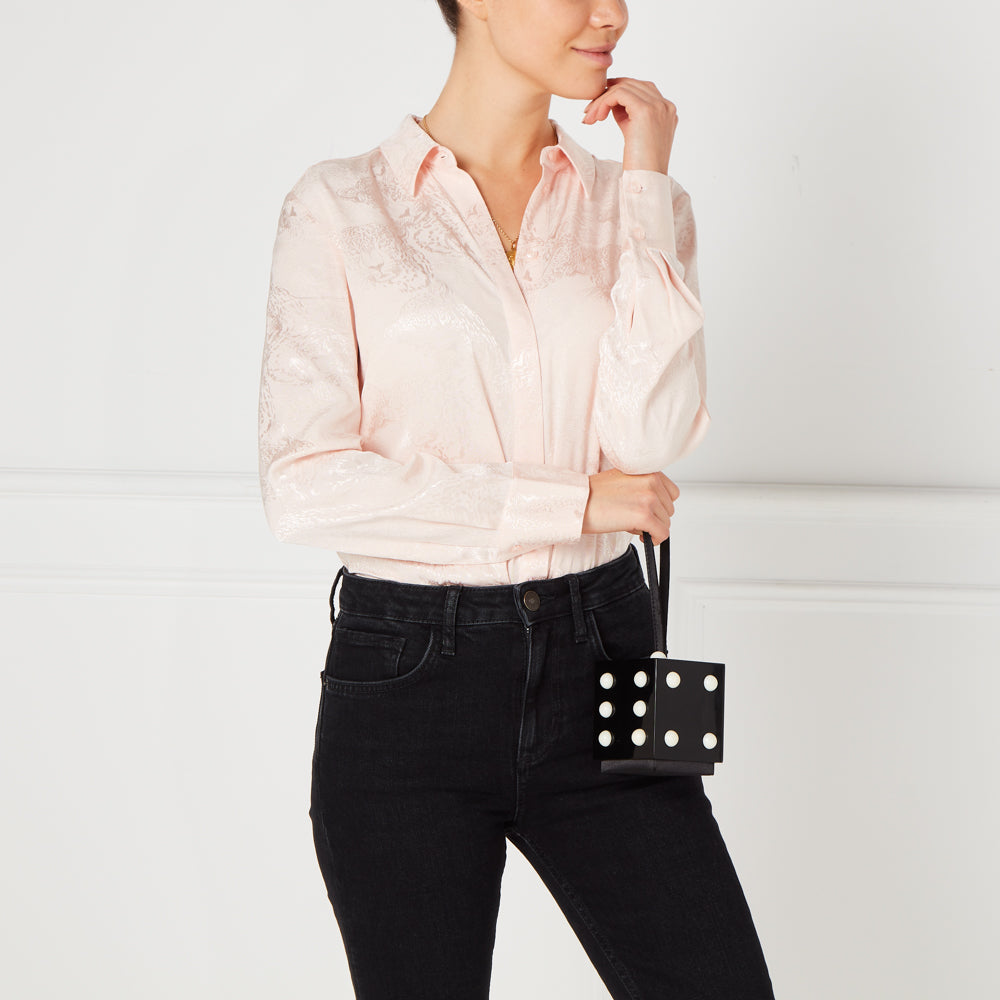 Woman in pink shirt and black jeans holding a black and white dice-shaped handbag against a white background.
