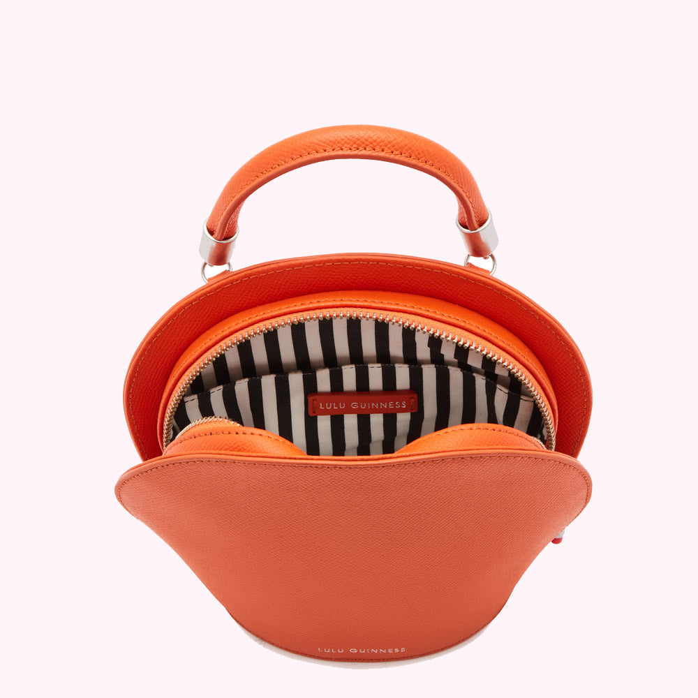 Overhead view of a bright orange Lulu Guinness handbag with a top handle and black and white striped interior.
