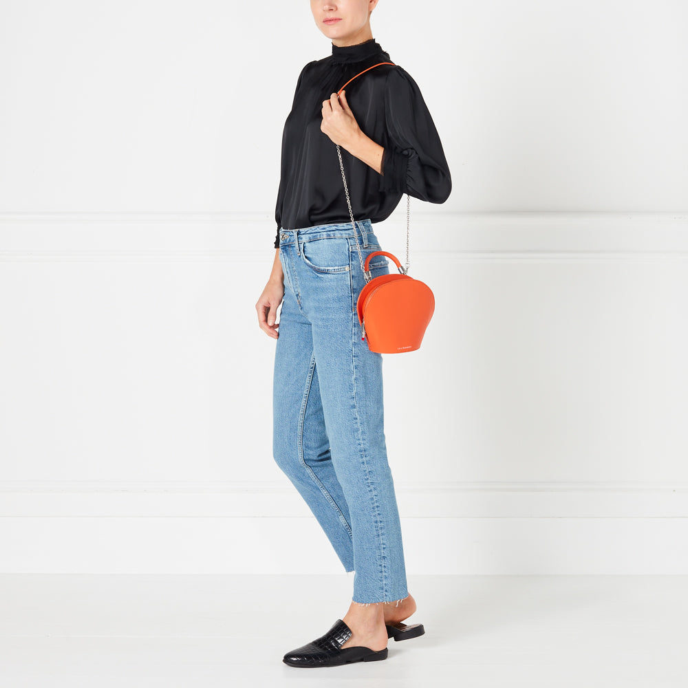 Woman wearing light blue jeans, black top, and black mules, carrying a bright orange round crossbody bag.

