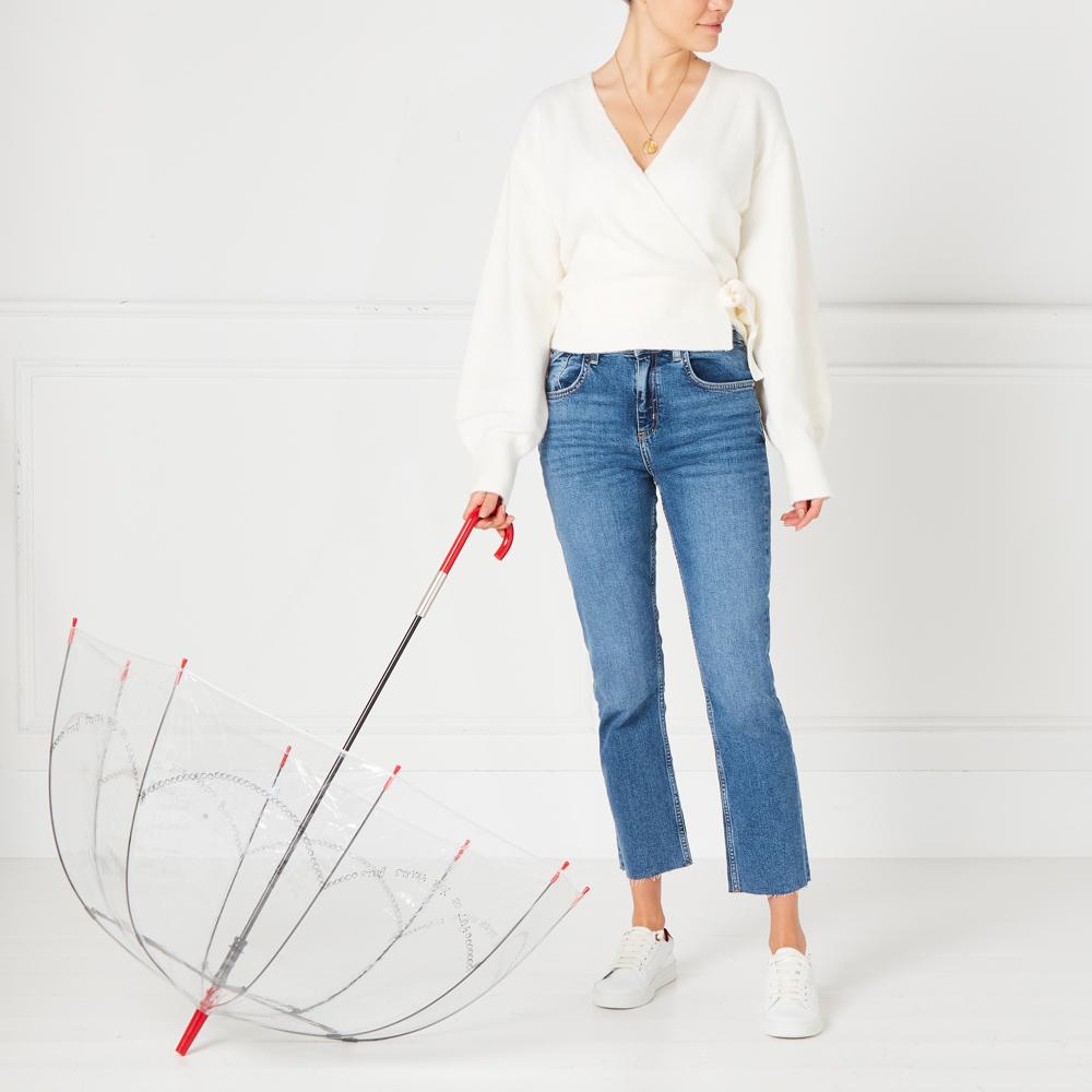 A woman in a white sweater and blue jeans holds a clear umbrella with a red handle against a white background.
