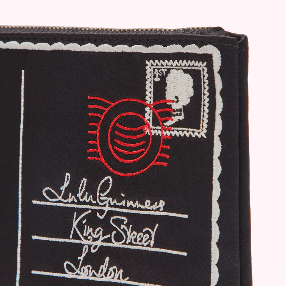 Close-up of a black pouch with embroidered postage stamp design, featuring red and white stitching. The address includes "Lulu Guinness," "King Street," and "London."
