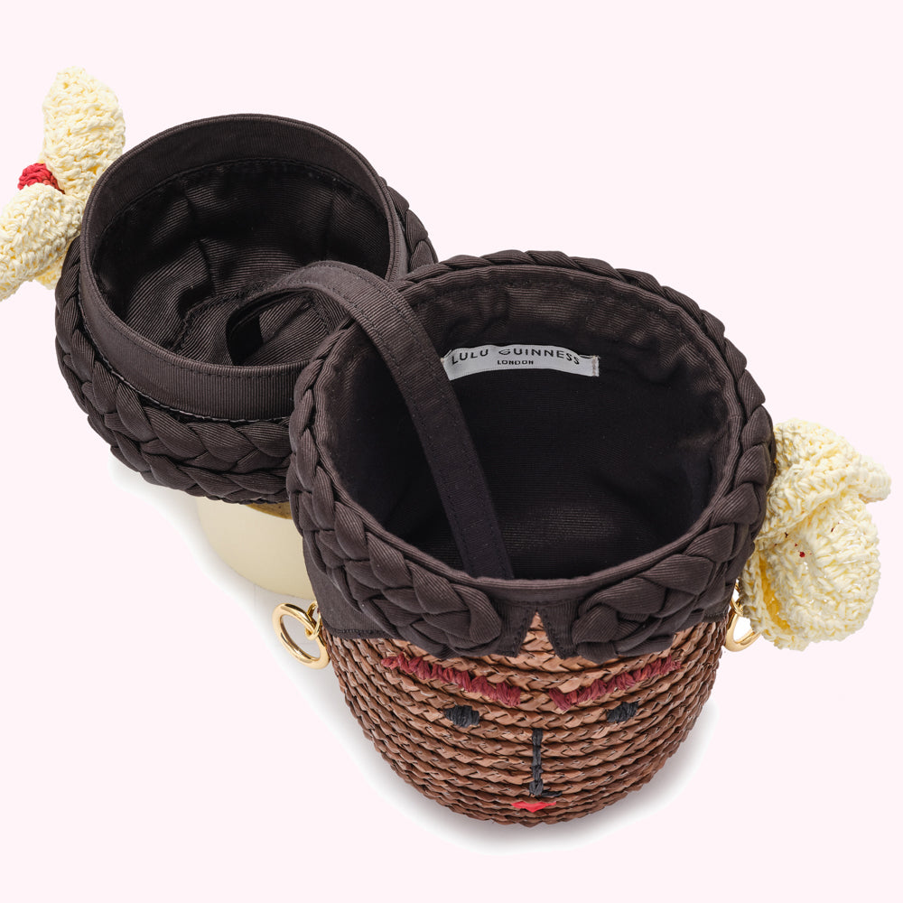 Overhead view of a Lulu Guinness London handbag, open to reveal its dark brown fabric interior; the handbag is designed in the shape of a woman's face with braided hair and features a woven straw exterior.