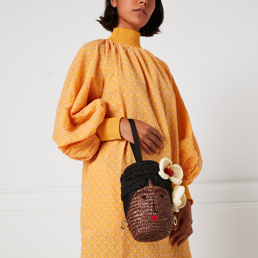 Woman in mustard yellow dress holding a woven handbag shaped like a woman's face with crocheted flowers.