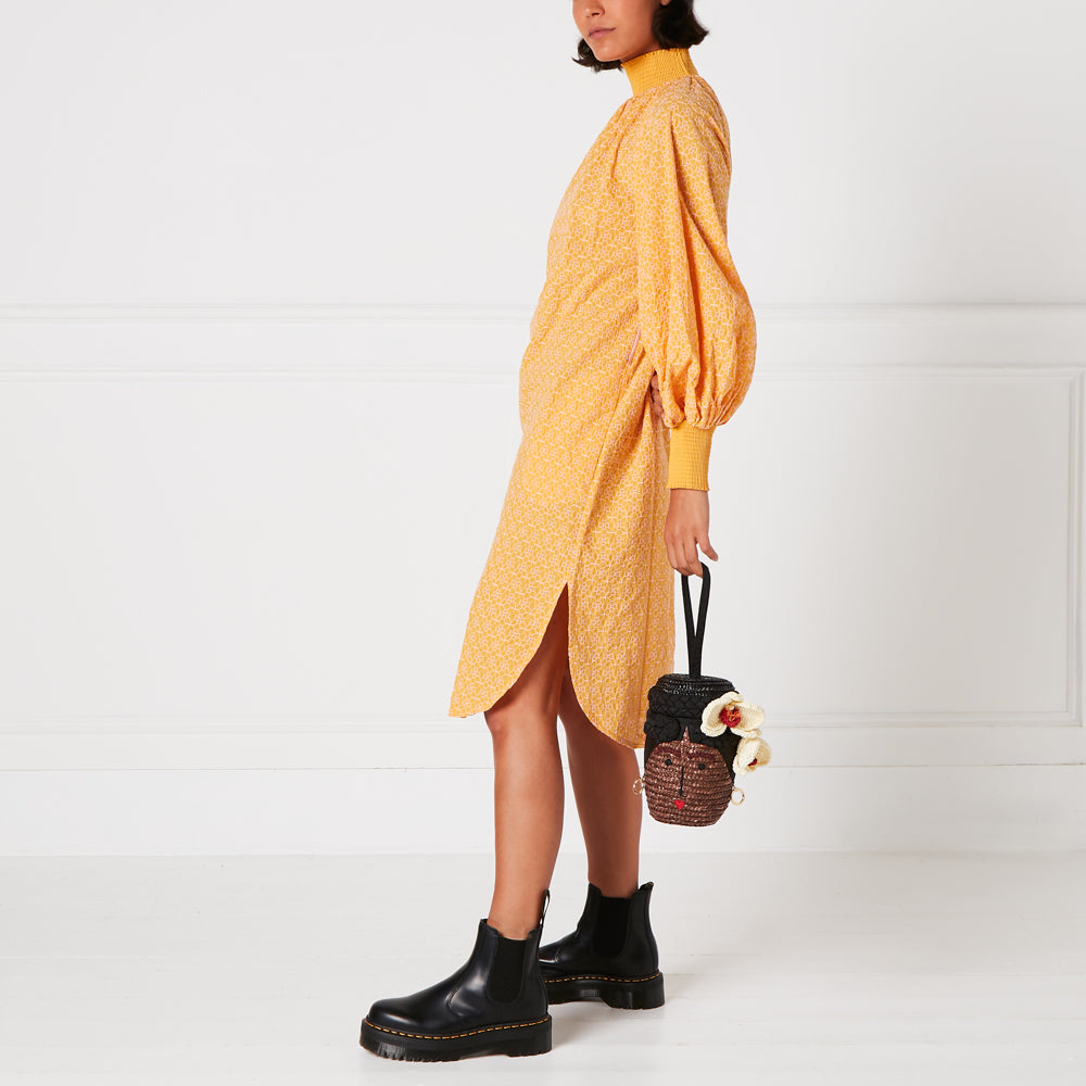 Woman in yellow floral dress and black boots holding a unique woven handbag with a face. The background is white.
