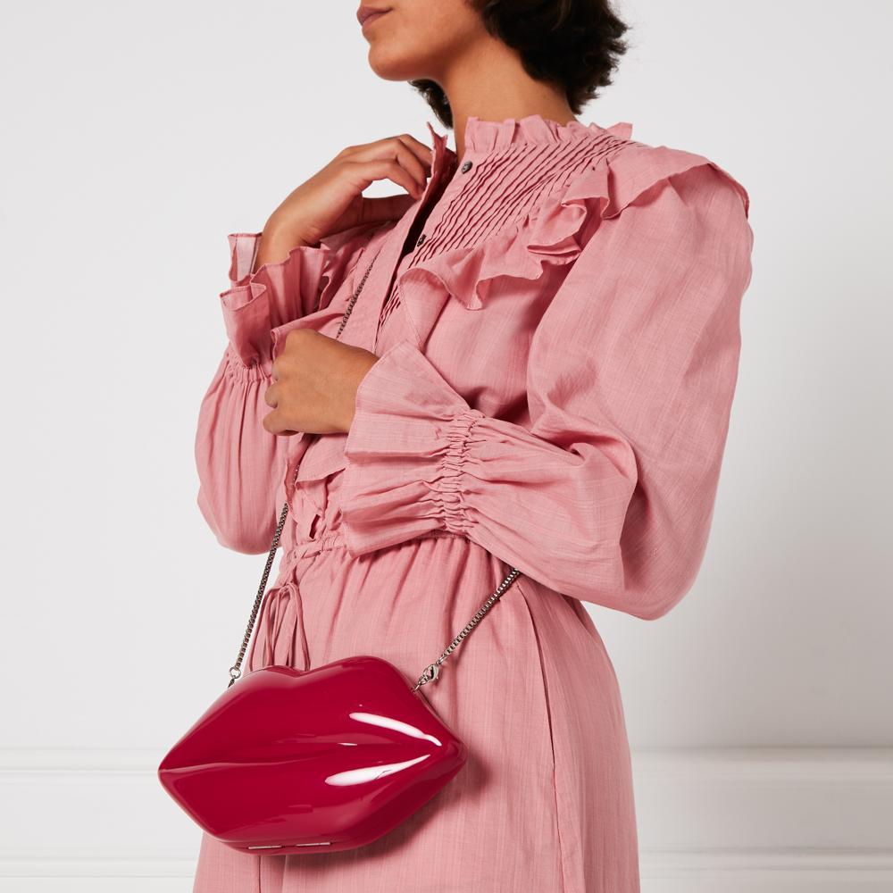 A woman in a pink dress holds a red lip-shaped crossbody bag.
