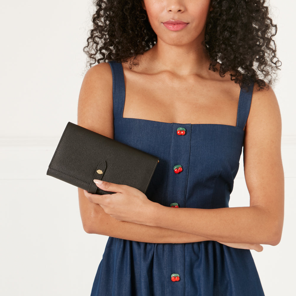 Woman with curly hair holding a black leather wallet in front of her. She is wearing a denim dress with cherry buttons.