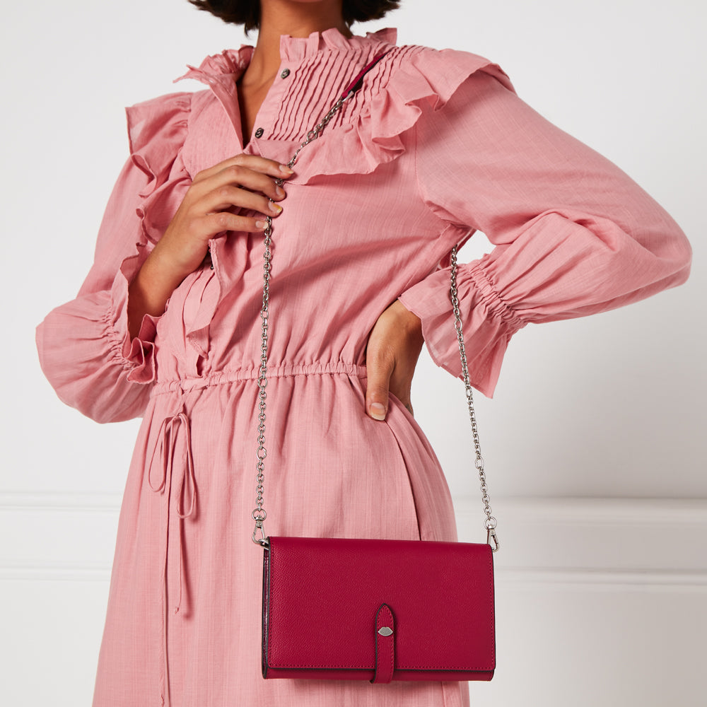 A crimson leather wallet-on-chain hangs from the shoulder of a model wearing a pink ruffled dress against a white background.
