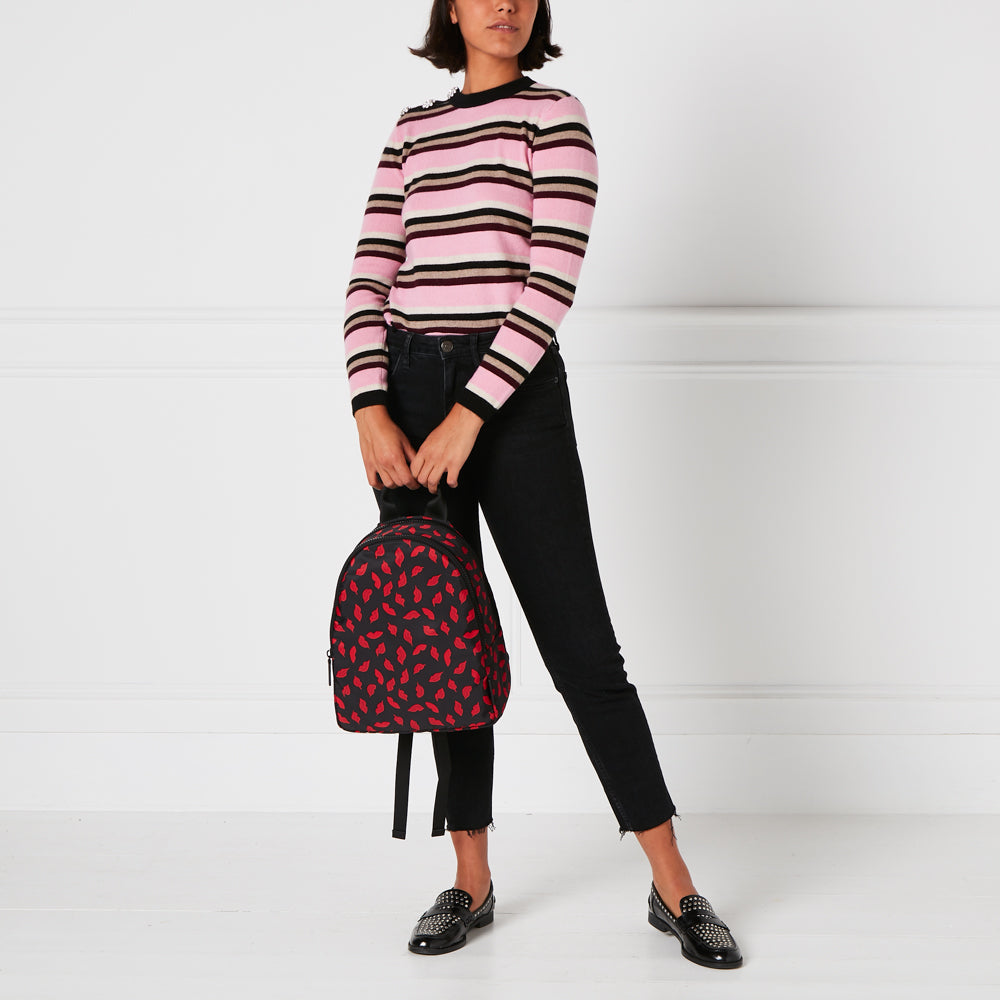 A person with short brown hair wearing a pink and brown striped sweater and black pants is standing in front of a white background. The person is holding a black backpack with a red lips pattern.