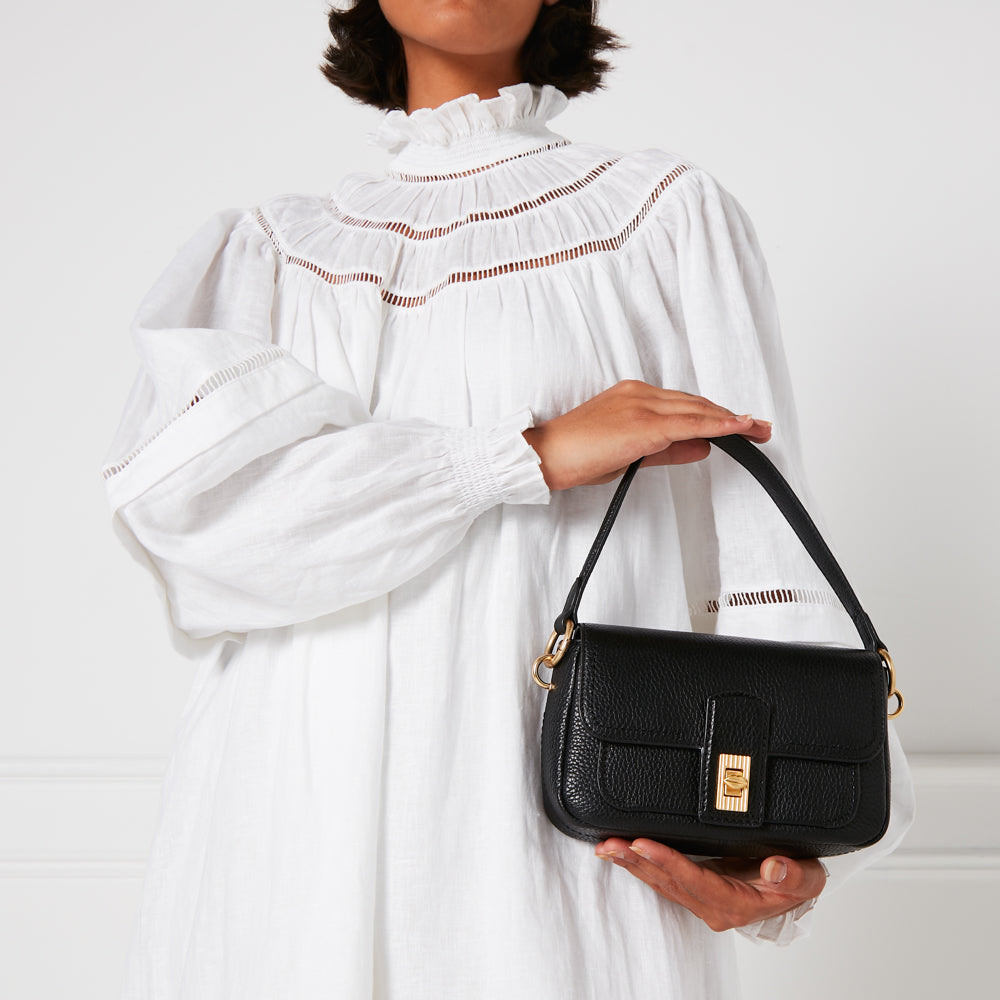 A woman in a white eyelet dress holds a small black handbag with gold hardware.
