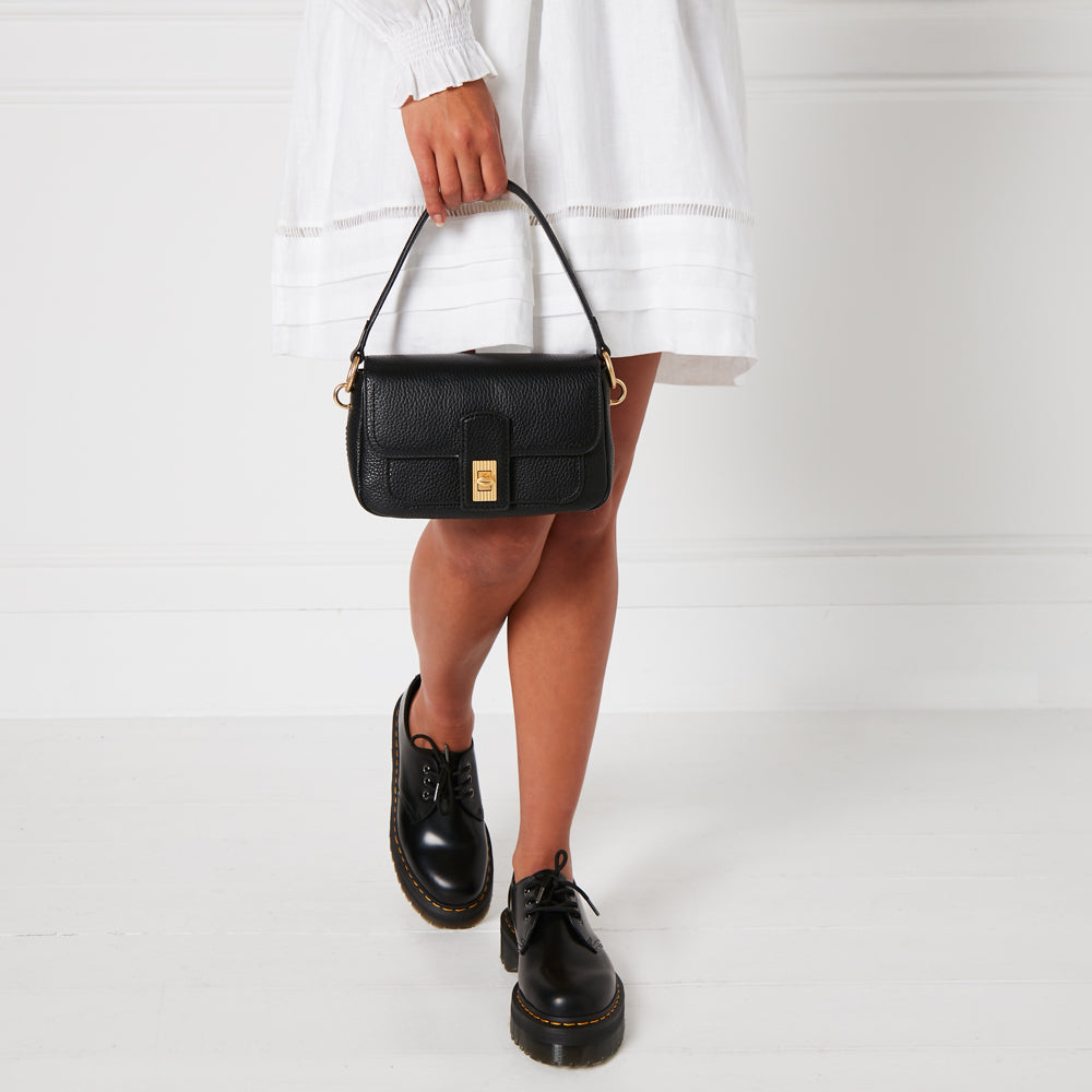 A woman in a white dress holds a black shoulder bag and wears black Dr. Martens shoes against a white background.