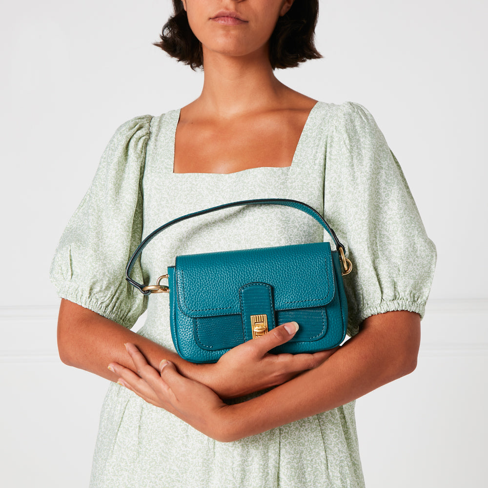 A teal leather shoulder bag held by a woman wearing a light green dress.
