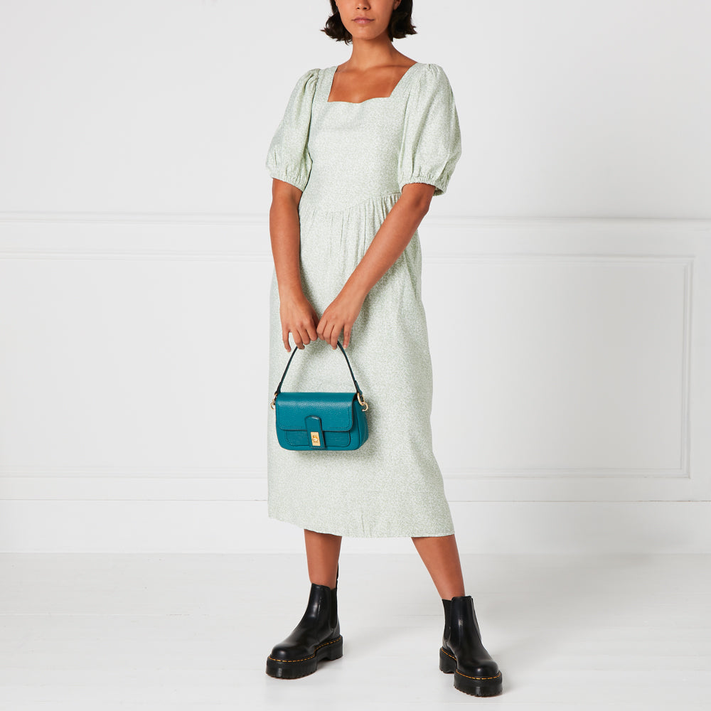 Woman in a light green dress holding a teal shoulder bag and wearing black Chelsea boots against a white background.
