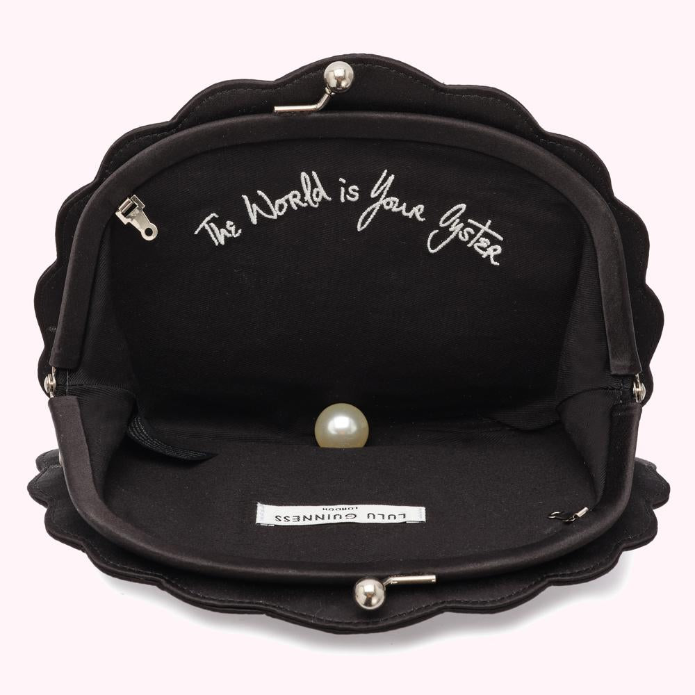 Open black Lulu Guinness shell-shaped clutch with the text "The world is your oyster" embroidered inside and a pearl resting on the bottom.
