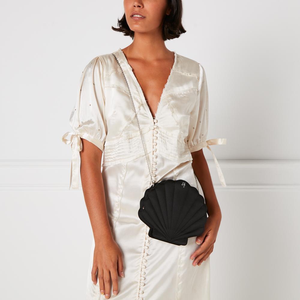 A woman in a cream-colored dress holds a black seashell-shaped crossbody bag against a white background.
