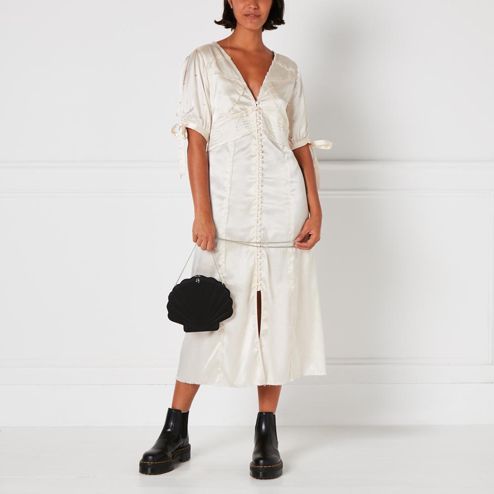 A woman in a cream midi dress and black ankle boots holds a black velvet shell-shaped handbag against a white background.
