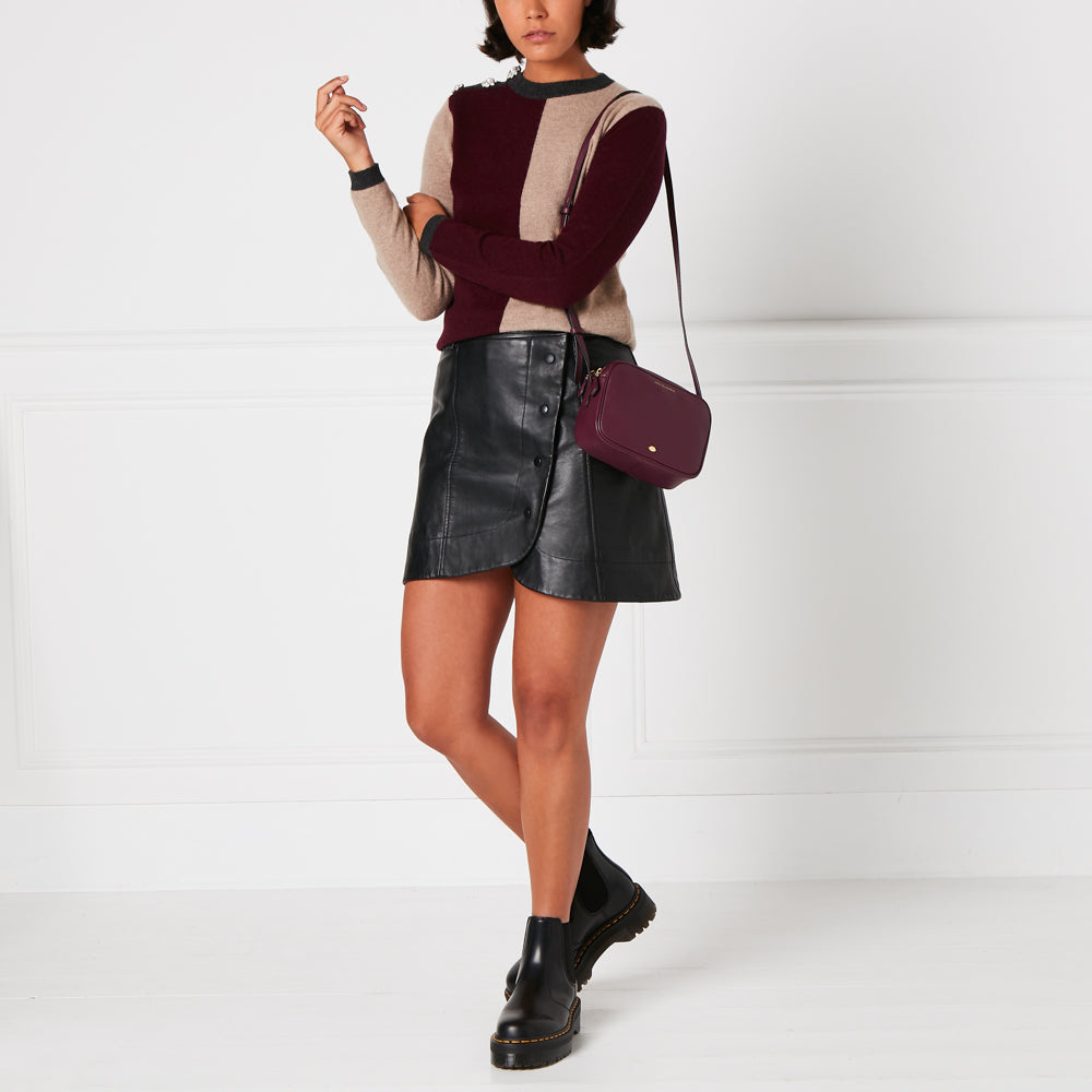 Woman wearing a burgundy and beige sweater, black leather mini skirt, and black Chelsea boots, carrying a dark burgundy crossbody bag against a white background.
