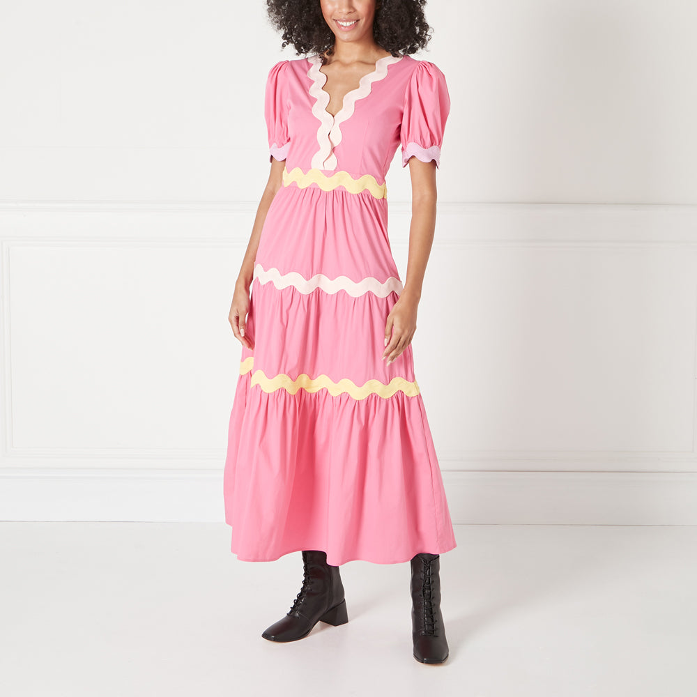 A model wears a pink tiered maxi dress with scalloped pastel yellow and lavender trim against a white background. She is wearing black lace-up ankle boots.
