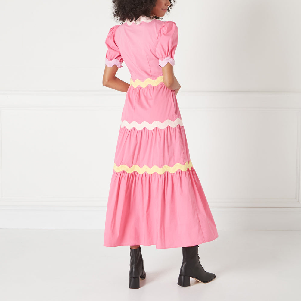 Rear view of a pink maxi dress with scalloped yellow and pink trim, worn by a model with dark hair.
