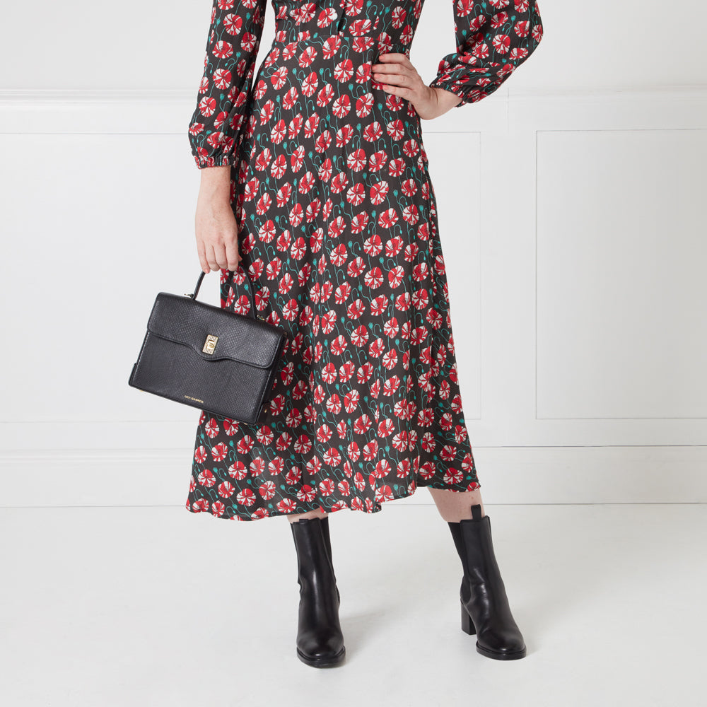 A woman in a floral midi dress and black ankle boots holds a black structured handbag.
