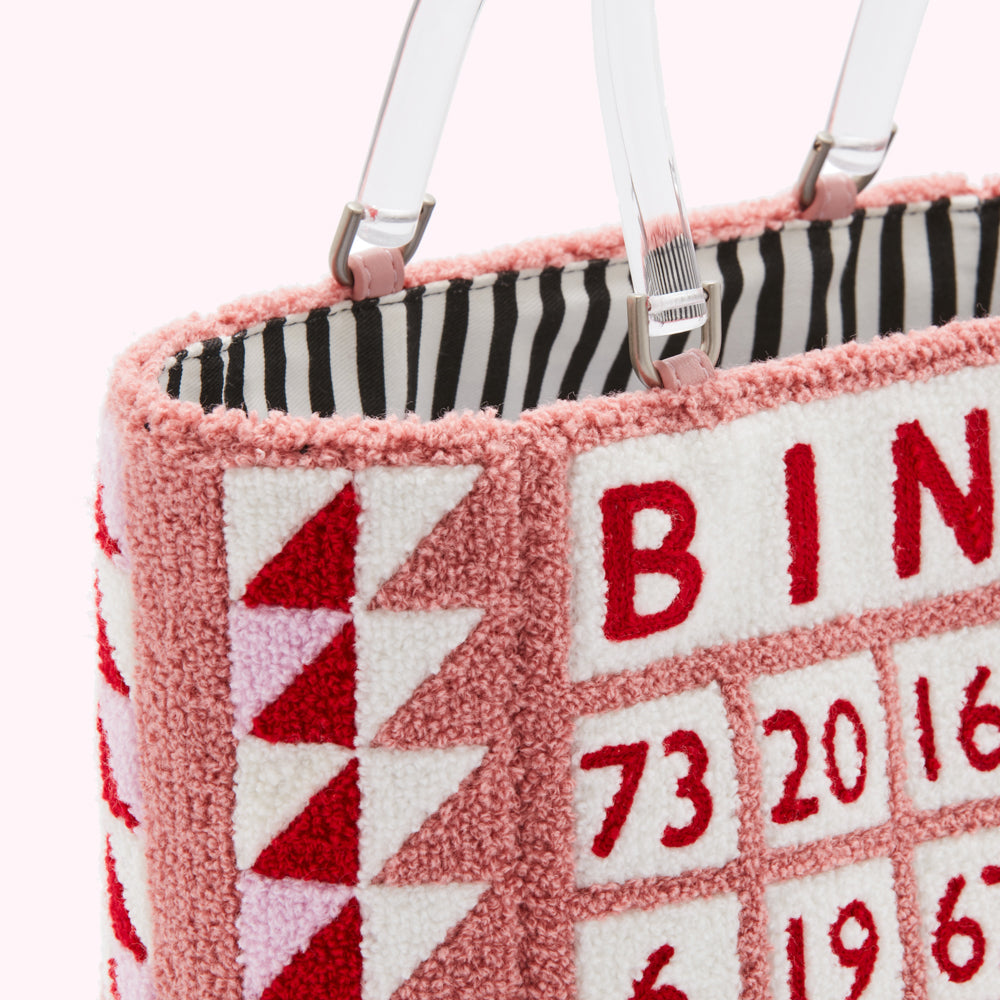 Close-up of a pink textured handbag with embroidered numbers and letters; the bag has clear handles and a black and white striped interior.
