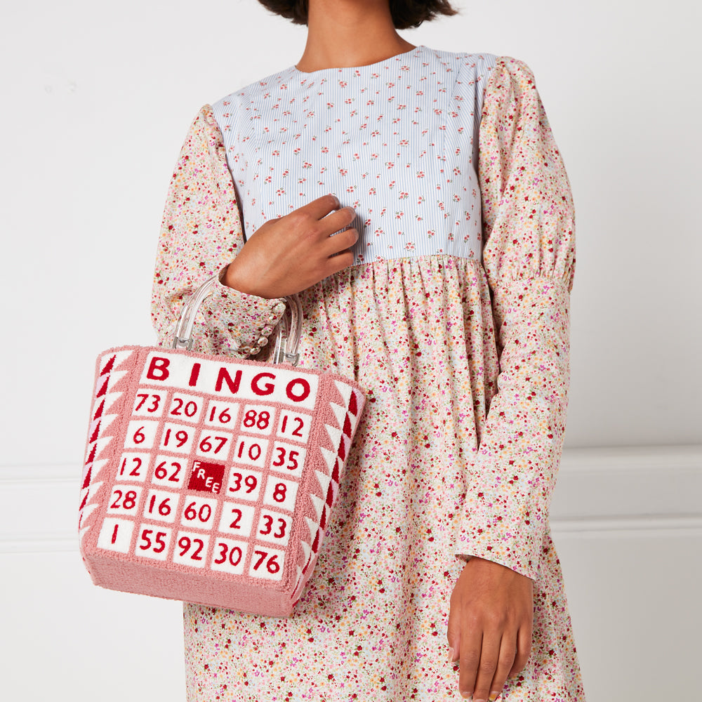 A pink bingo-themed tote bag with a clear handle is held by a person wearing a floral dress.