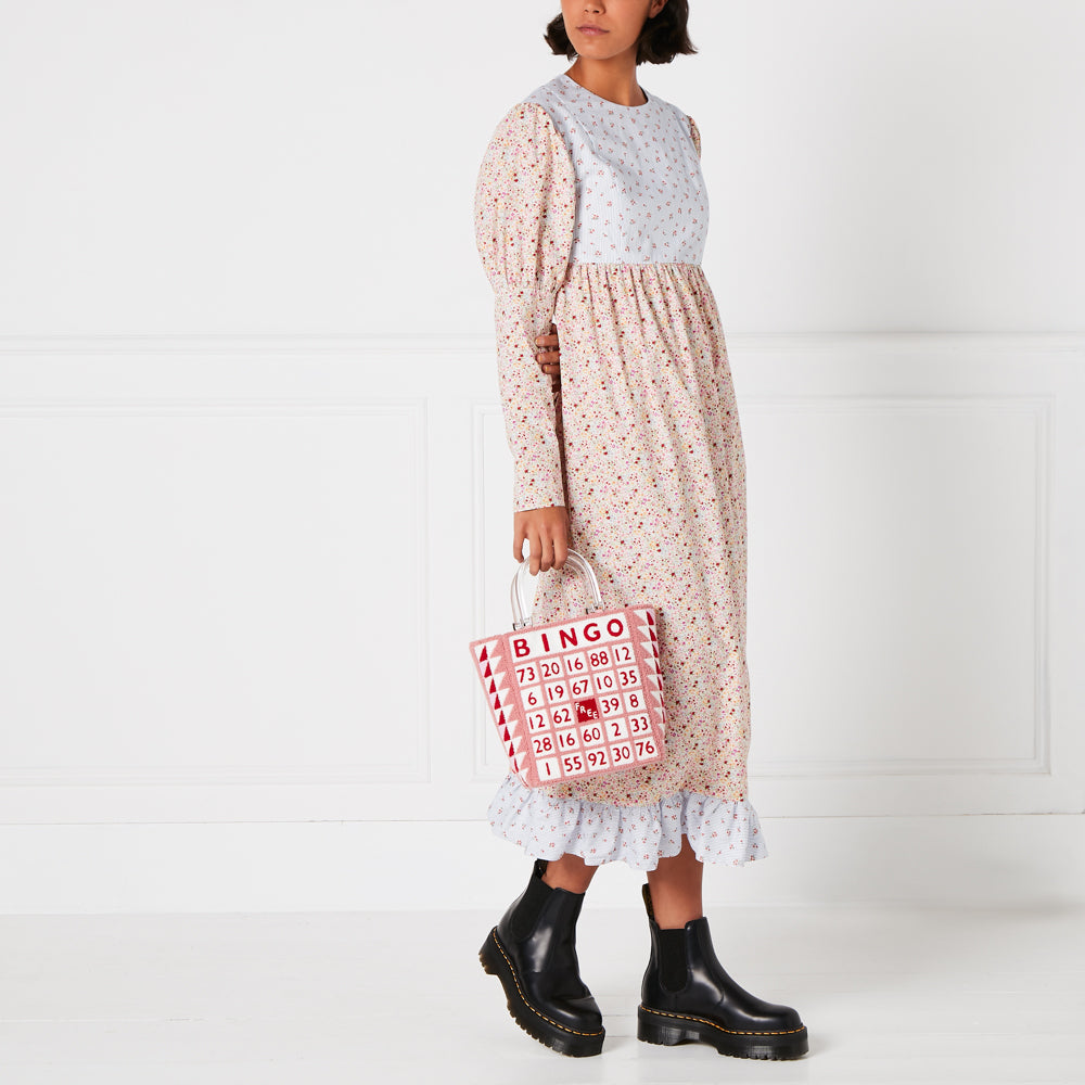 A person wearing a floral dress and black boots holds a pink bingo-themed tote bag against a white background.