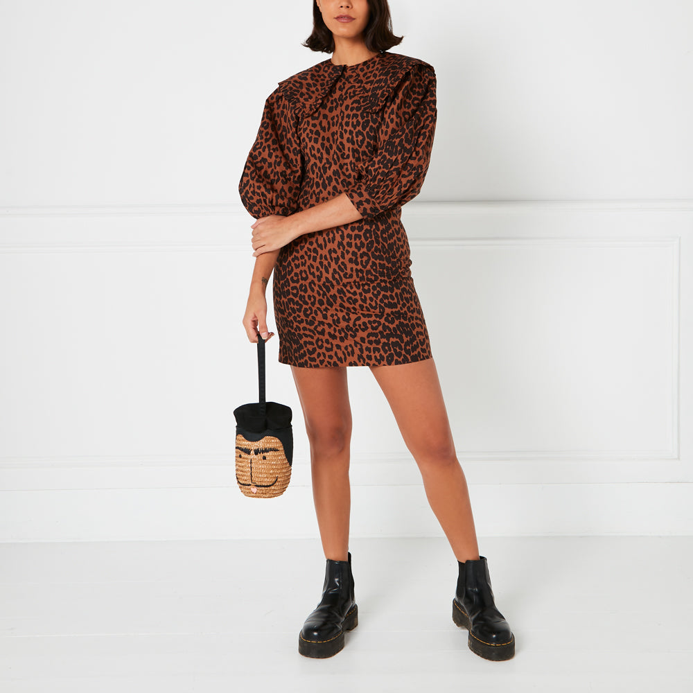 A model wears a brown and black leopard print mini dress with a large collar and black Chelsea boots. She carries a straw bag shaped like a face. The background is white.
