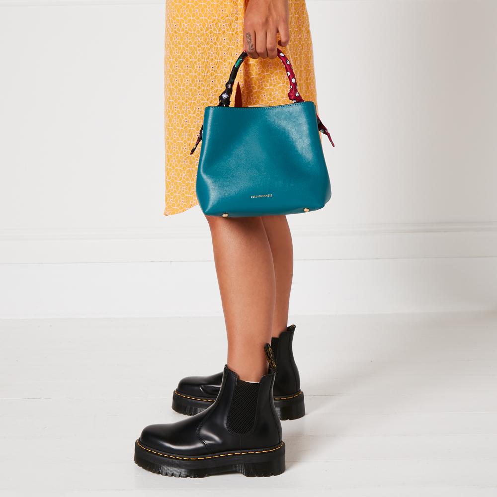 A teal Lulu Guinness handbag with a colourful scarf-print handle held by a person wearing black Dr. Martens boots and a yellow dress against a white background.
