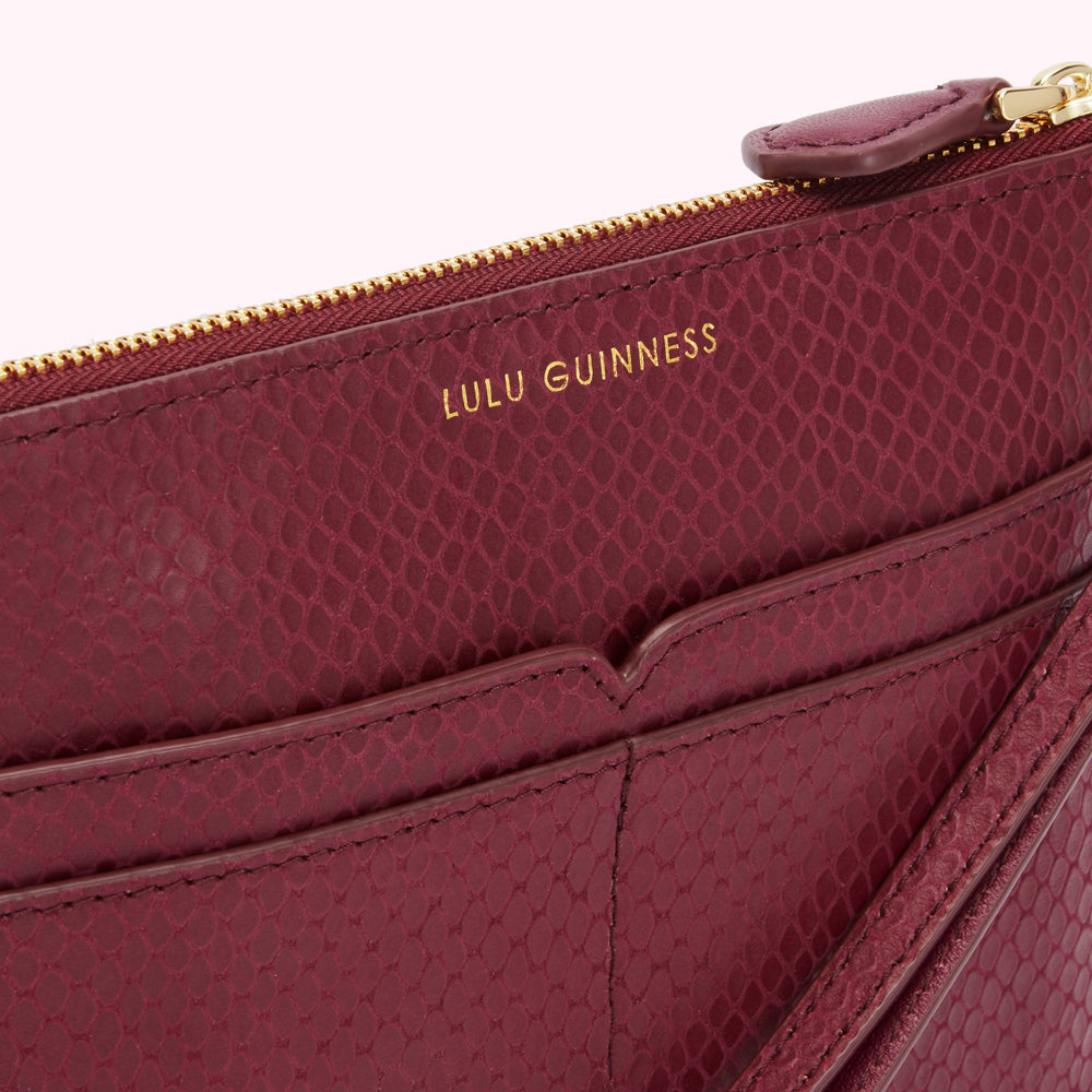 Close-up of a burgundy Lulu Guinness snakeskin-textured zip pouch with a gold zipper and strap.
