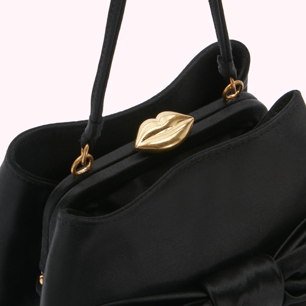 Close-up of a black satin handbag with a gold lip-shaped clasp and a bow detail.
