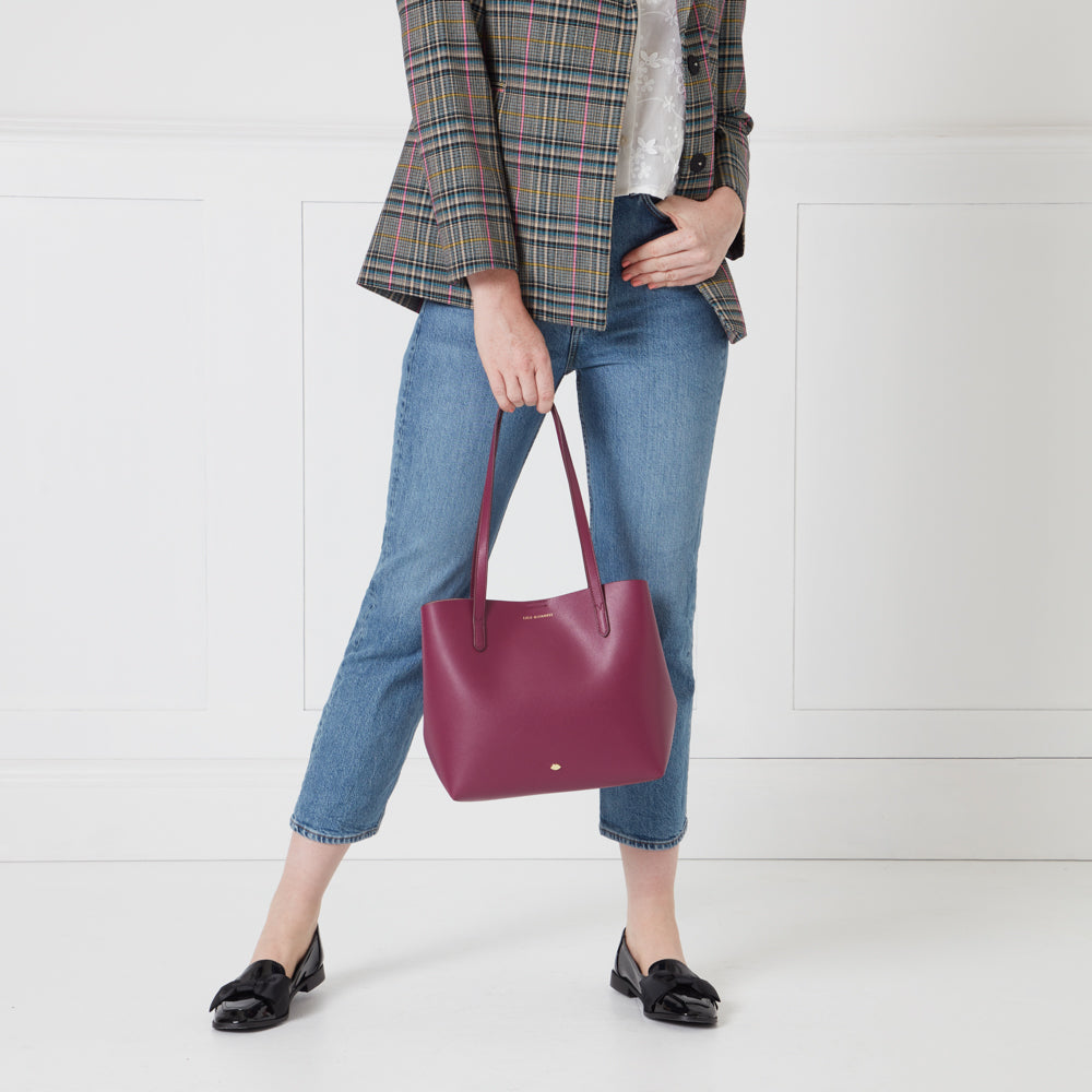 A woman in light blue jeans and a checked blazer holds a burgundy tote bag.
