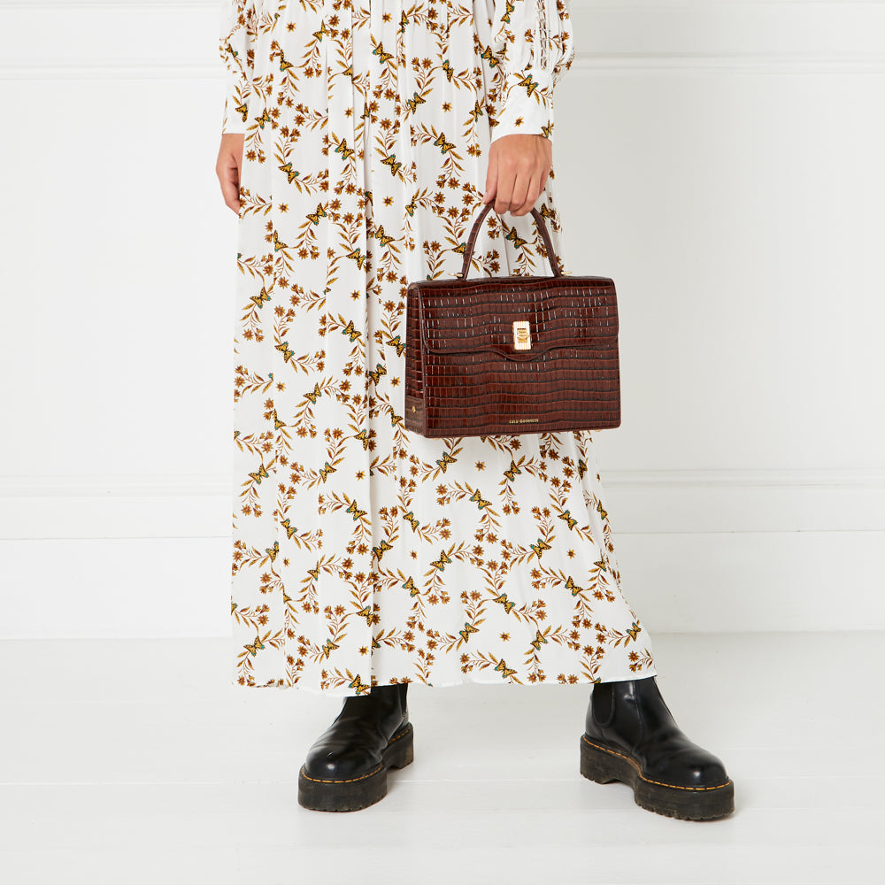 A woman in a white maxi dress with a brown butterfly pattern holds a brown crocodile-embossed handbag.
