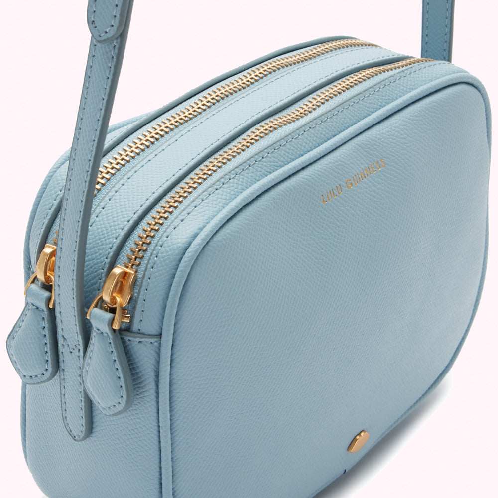 Close-up of a pale blue leather Lily Guinness crossbody bag with two gold zippers.