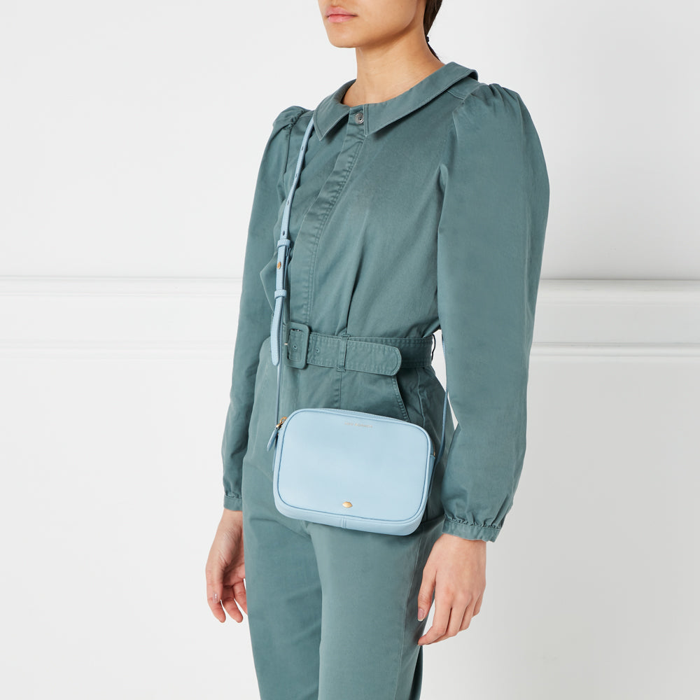 A pale blue crossbody bag worn by a model in a sage green jumpsuit against a white background.

