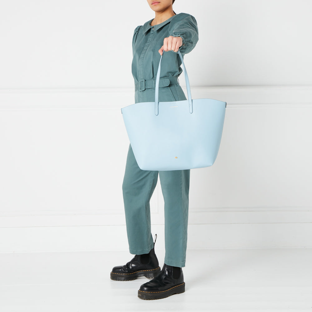A person in teal pants and black boots holds a light blue tote bag against a white background.
