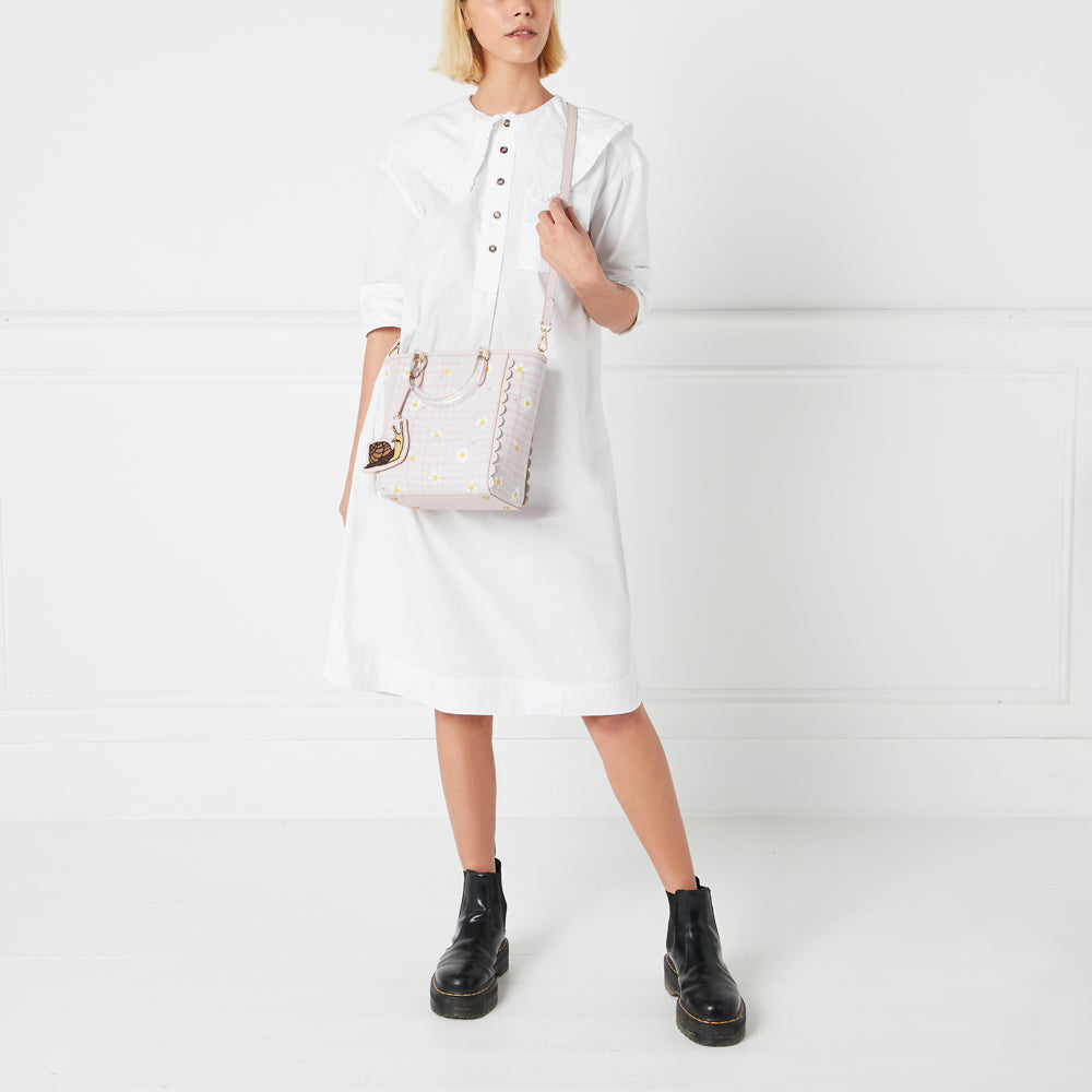 A woman in a white dress holds a pale pink daisy-printed tote bag with a snail charm.
