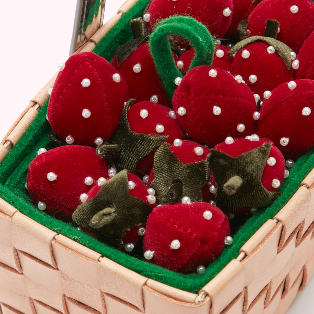 Close-up of a beige woven basket filled with red velvet strawberries adorned with white pearls. The strawberries have green velvet leaves.
