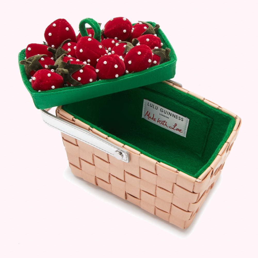 Pink woven basket-shaped purse with a removable lid containing a cluster of red velvet strawberries; the interior is lined in green. The label reads "Lulu Guinness London, Made with Love."
