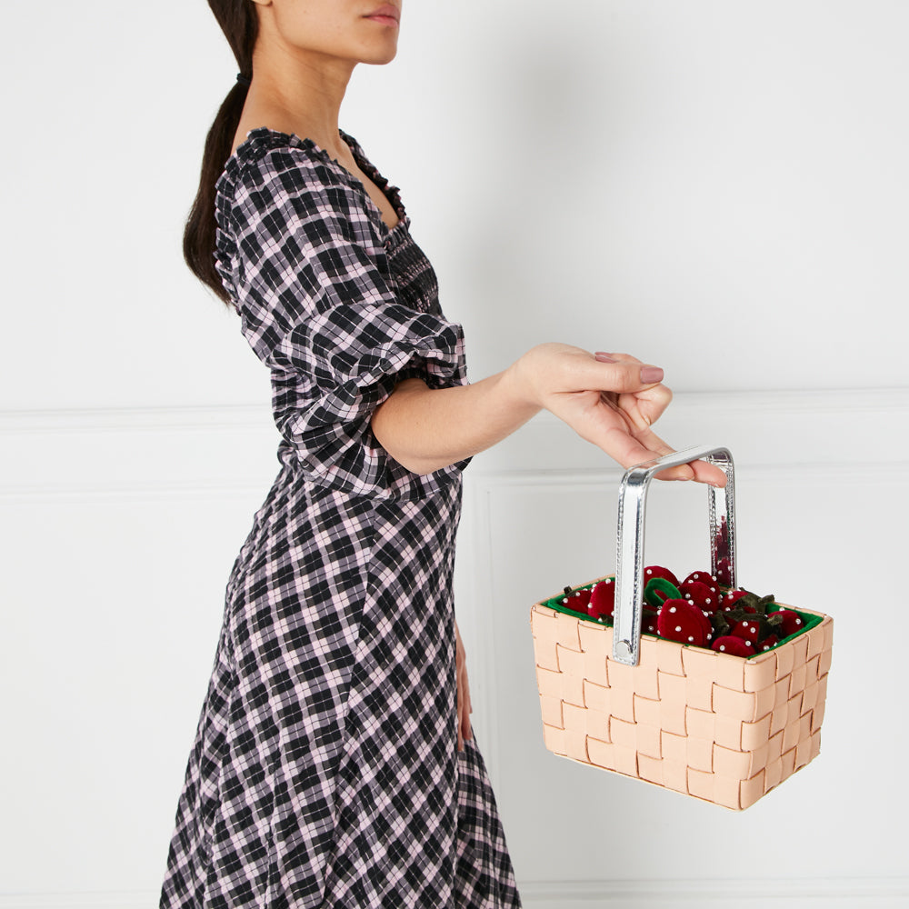 Woman in black and pink gingham dress holding a beige woven basket bag filled with red strawberries against a white background.
