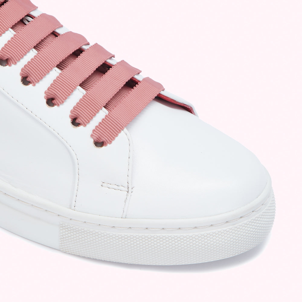 Close-up of white leather sneakers with dusty rose grosgrain ribbon laces against a pink background.
